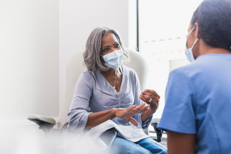 A senior woman, wearing a protective face mask, talks with a female nurse during a medical appointment